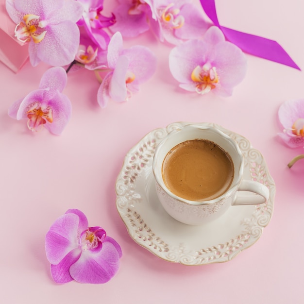 Composición delicada flatlay con taza de café matutino con leche o capuchino, bolsa de regalo rosa y flores de orquídeas sobre fondo rosa claro