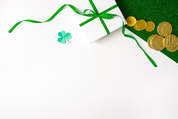 composición del día de San Patricio. Caja de regalo blanca con un lazo verde, un trébol de papel y monedas de oro sobre un fondo verde brillante y blanco