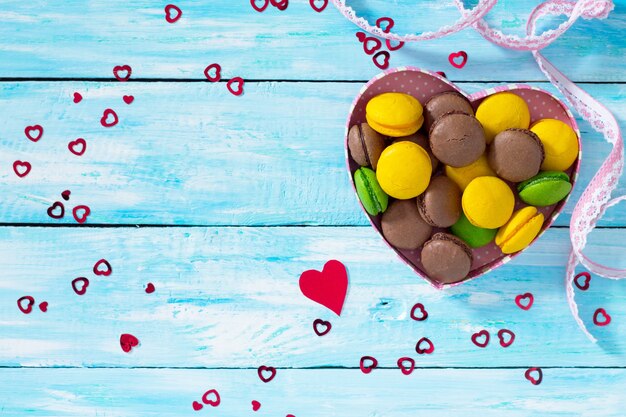 Composición del día de San Valentín con macarrones en recipiente en forma de corazón sobre una mesa de madera azul. Vista superior.