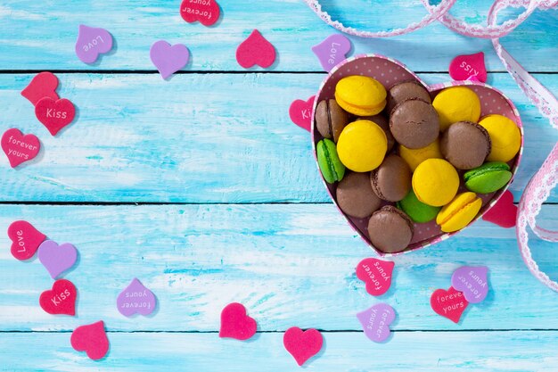 Composición del día de San Valentín con macarrones en recipiente en forma de corazón sobre una mesa de madera azul. Vista superior.