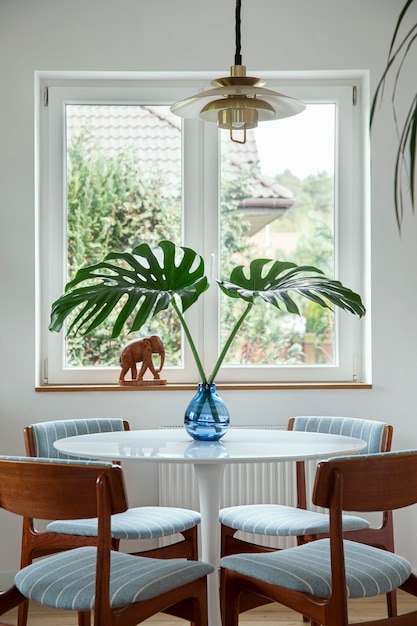 Composición elegante del interior del comedor con mesa de diseño, sillas, hojas tropicales en jarrón, ventana y decoración elegante en la decoración del hogar. Plantilla.