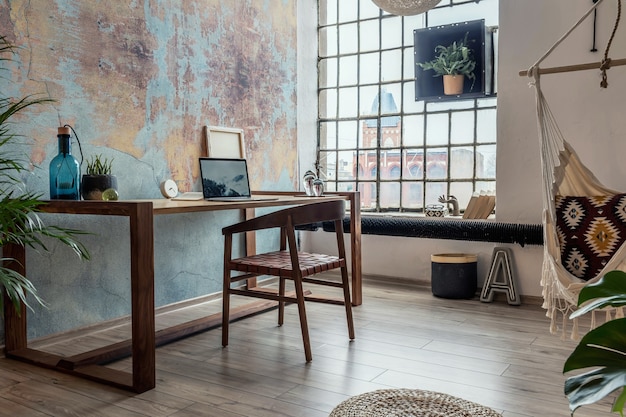 Composición elegante y moderna del interior del espacio de trabajo creativo con mesa y silla de madera, plantas y accesorios. Amplia habitación con paredes creativas y suelo de parquet.