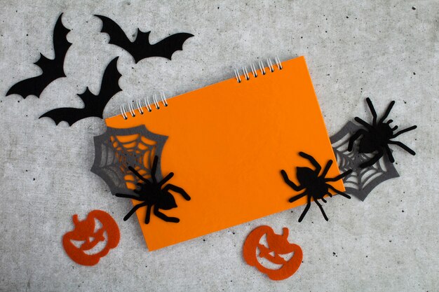 Composición para Halloween con cuaderno naranja, arañas decorativas, calabazas, telas de araña y murciélagos sobre fondo gris. Copie el espacio. Vista superior.