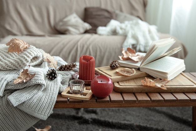 Una composición hogareña acogedora con velas, un libro, suéteres de punto y hojas en el interior de la habitación.