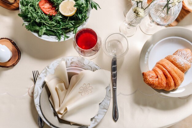 Composición laica plana con una mesa puesta. Un plato con una servilleta, una copa de vino, un croissant y una ensalada en la mesa.