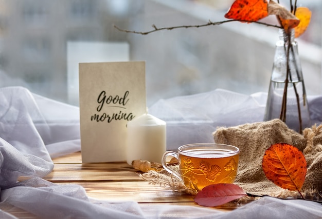 Composición de la mañana con una taza de té de vidrio transparente, un jarrón y una tarjeta "Buenos días". Concepto de una sensación de comodidad en casa. Enfoque selectivo.
