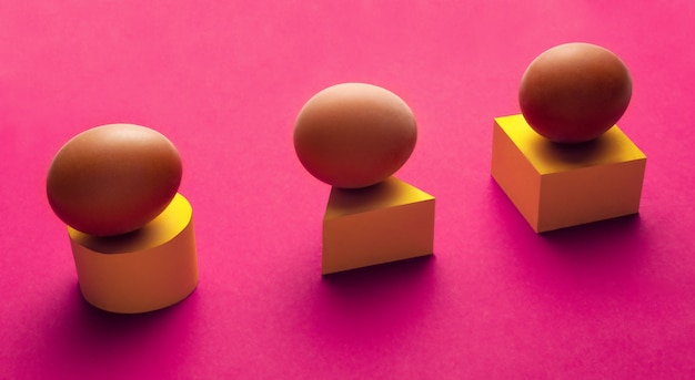 Composición mínima creativa de tres huevos sobre un cilindro amarillo, triángulo y cubo sobre una superficie violeta.