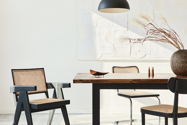 Composición minimalista del interior del comedor con mesa de madera, sillas de diseño, flores secas en un jarrón, lámpara colgante negra, cuadros de arte en la pared y elegantes accesorios personales