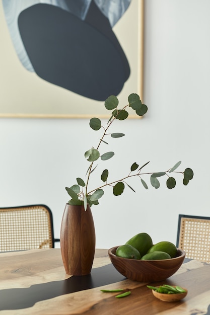 Composición minimalista en la mesa de madera de diseño con frutas, hojas tropicales en jarrón, pinturas abstractas y elegante silla