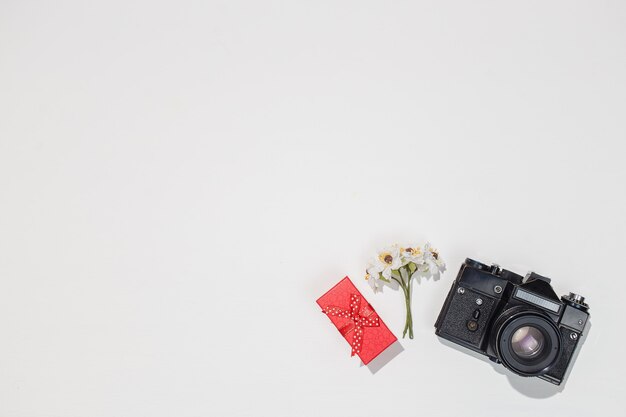 Composición minimalista en plano con cámara retro, caja de regalo roja y flor de campo de primavera sobre fondo blanco.