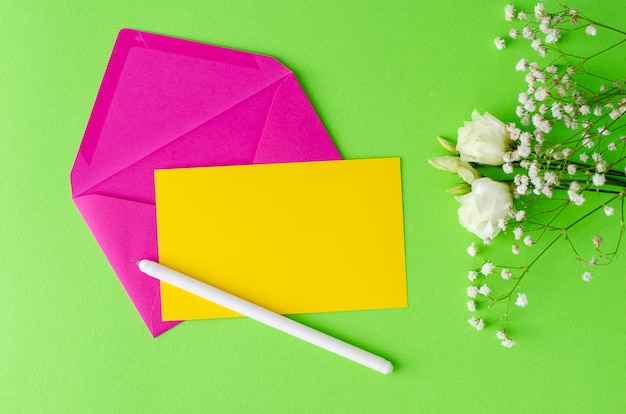 Composición minimalista con un sobre rosa, tarjeta amarilla en blanco, pluma y flores. Flay laico, concepto de maqueta.