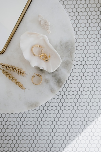 Composición de moda mínima con aretes dorados en concha sobre mesa de mármol con espejo y tallos de trigo