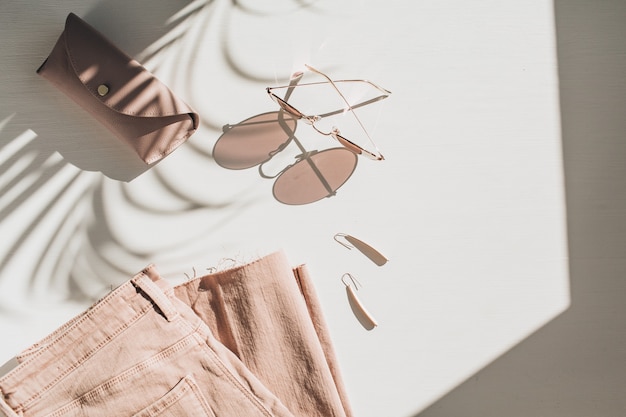 Composición de moda con ropa de mujer y accesorios en blanco. Pendientes, gafas de sol, culottes de jean rosa sobre blanco