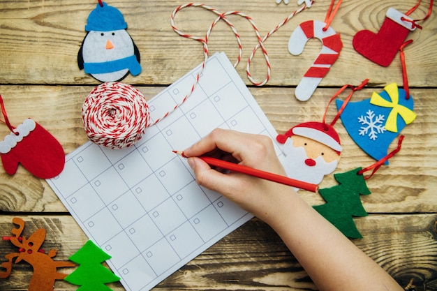 Foto una composición navideña con un planeador o calendario la mano de una mujer sosteniendo un lápiz juguetes hechos a mano de fieltro plano mitones rojos sentían el reno de santa árbol de navidad sobre un fondo de madera