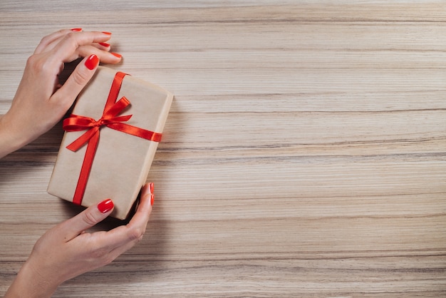 Foto composición navideña. vista superior, endecha plana. manos femeninas con caja de regalo envuelta en papel artesanal y decorada con cinta de raso roja sobre mesa de madera