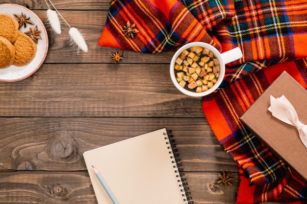 Composición de otoño. Bufanda roja de moda para mujer, libreta de papel, taza de té de hierbas, flores secas, galletas, caja de regalo