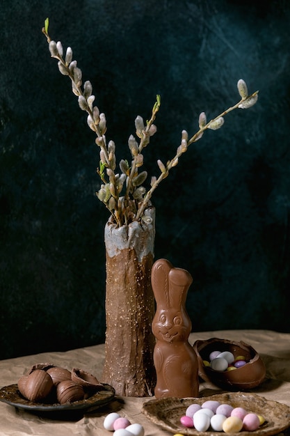 Composición de Pascua con ramas de sauce en flor en florero de cerámica, conejo de chocolate tradicional, huevos y dulces en la mesa con papel artesanal arrugado