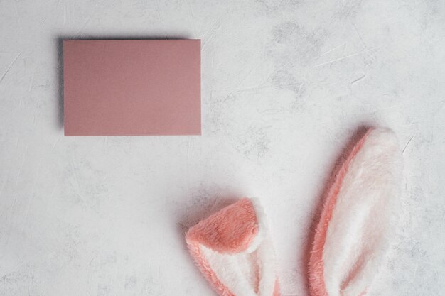 Composición de Pascua de tarjeta de papel de felicitación vacía de poste rosa sobre fondo de papel gris blanco y corte de orejas de pelo esponjoso de juguete de conejito Celebración de fiesta tradicional Lugar para texto