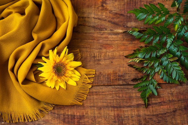 Composición plana con bufanda de punto amarilla, ramita verde y girasol sobre una mesa de madera