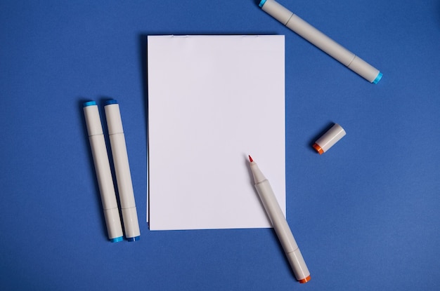 Composición plana de dos colores con marcadores de acuarela o rotuladores con hoja de papel en blanco con espacio de copia aislado sobre fondo azul.