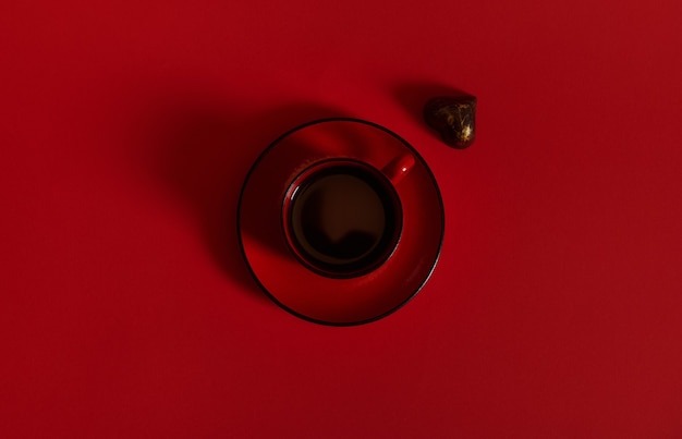 Composición plana con una taza de café roja sobre un platillo de cerámica y un praliné en forma de corazón sobre una superficie roja. Concepto de día del chocolate sobre fondo rojo con espacio de copia