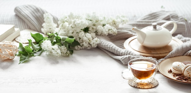 Composición de primavera acogedora con una taza de té, una tetera, macarrones franceses, color lila sobre una mesa de luz borrosa.