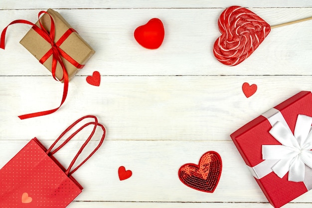 Composición romántica creativa del día de San Valentín con corazones rojos, piruleta, caja de regalo y bolsa de papel sobre fondo blanco.