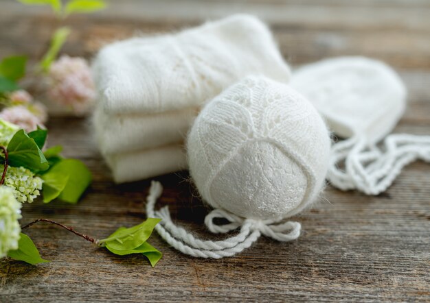 Composición de ropa blanca tejida para recién nacido en la mesa de madera con flores. Conjunto de diseño de ropa de lana infantil con sombrero.