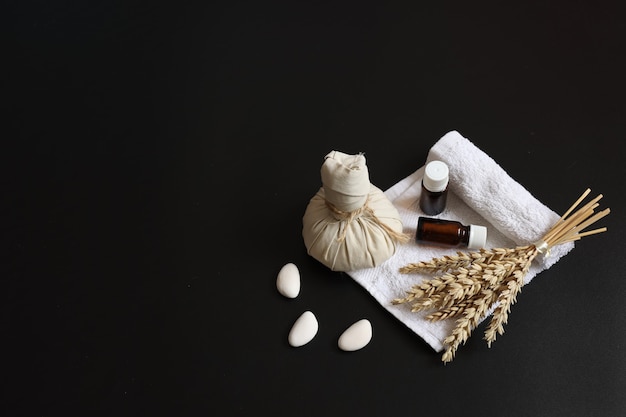 Composición de spa con bolsa de masaje a base de hierbas, ramita de trigo, aceites y esponja vegetal sobre fondo negro, espacio de copia.