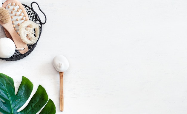 Composición de spa con cepillo de masaje y otros accesorios de baño en canasta sobre fondo blanco con hoja de monstera.