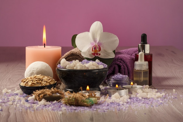 Composición de spa con flor de orquídea, cuenco con sal marina, botellas con aceite aromático, jabón, exfoliante, velas y toallas sobre tabla de madera y fondo rosa