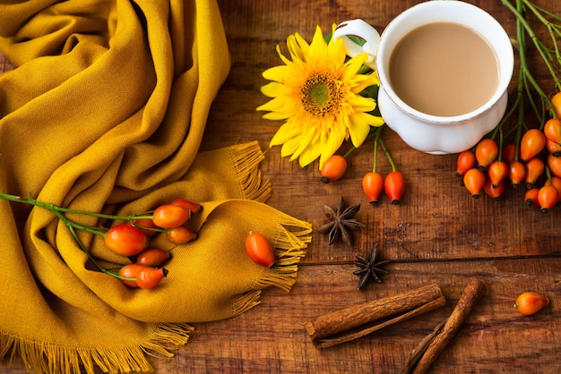 Composición de la taza de té de otoño, pañuelo amarillo, bayas de rosa y flor de girasol sobre un fondo de madera