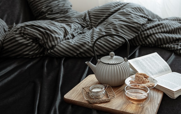 Composición con té en una tetera, galletas, un libro y una vela en la cama