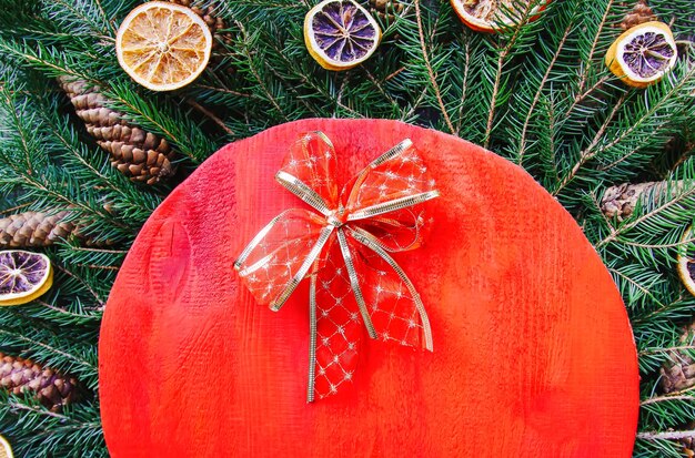 Composición de vacaciones de invierno de Navidad y año nuevo con superficie de madera roja sobre ramas de abeto verde, rodajas de naranja secas, conos, caja de regalo y lazo rojo.