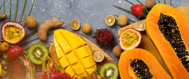 Composición de verano de papaya en rodajas por la mitad, fruta del dragón, rambután, tamarindo, mango, maracuyá, kiwi, fresa, hojas. Endecha plana. Vista superior de frutas tropicales sobre fondo azul grisáceo.