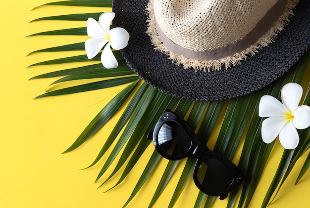 Composición de verano con sombrero, gafas de sol, hojas de palmeras tropicales y flores sobre fondo amarillo
