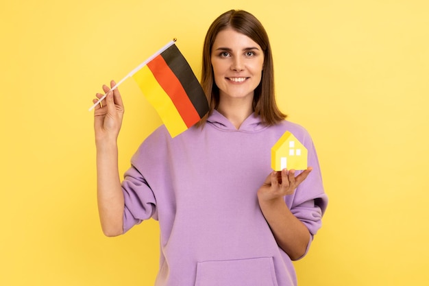 Compra de bienes raíces en la inmigración de Alemania Retrato de una mujer positiva sosteniendo una bandera alemana y una casa de papel con una sudadera con capucha púrpura Disparo de estudio interior aislado sobre fondo amarillo