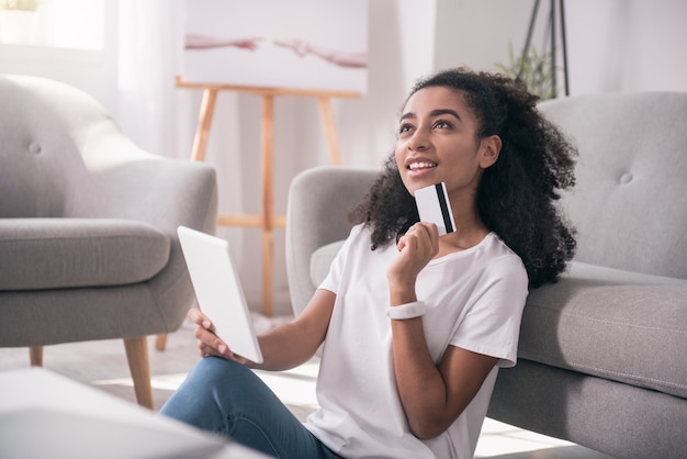 Compra pela Internet. Mulher bonita e alegre segurando o cartão de crédito enquanto pensa no que comprar na Internet