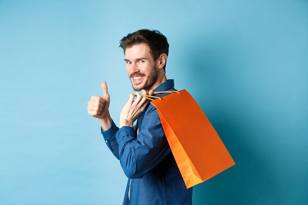 Comprador alegre que sostiene la bolsa de compras naranja en el hombro, da la vuelta a la cámara con los pulgares hacia arriba, recomendando la tienda, fondo azul.