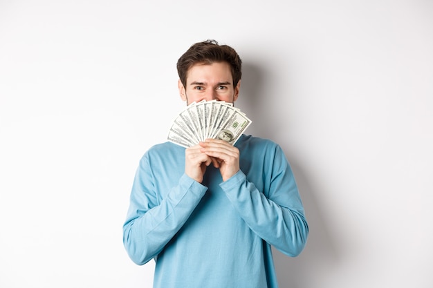 Comprador alegre segurando dinheiro para fazer compras, em pé com dólares e sorrindo, em pé sobre um fundo branco em roupas casuais.