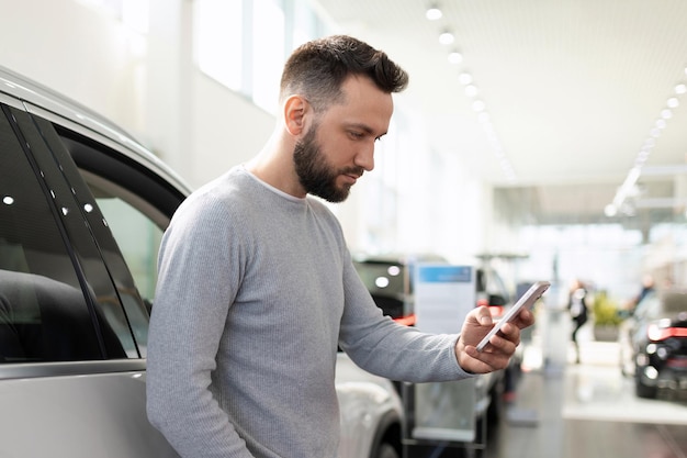 El comprador en el centro de automóviles del concesionario elige un automóvil compara los precios en un teléfono inteligente a través del