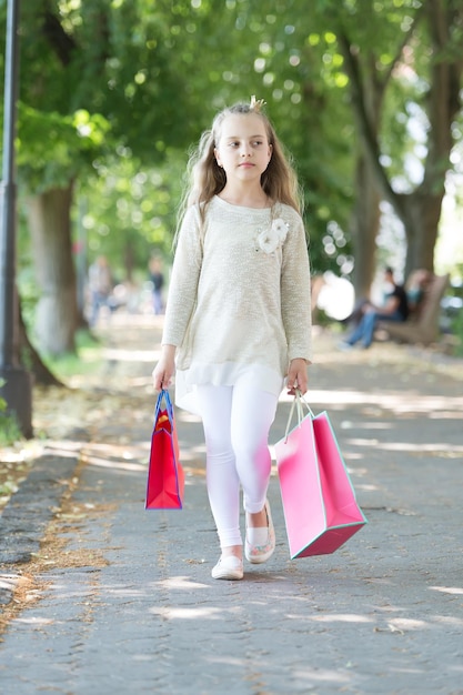 Comprador de criança em roupas de moda ao ar livre Criança com sacos de papel no verão Pequena princesa com coroa em longos cabelos loiros Compras à venda e compra Menina com sacolas de compras passear no parque