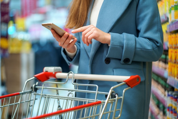 Comprador de mulher com carrinho no corredor da loja com lista de compras em smartphone durante a compra de alimentos