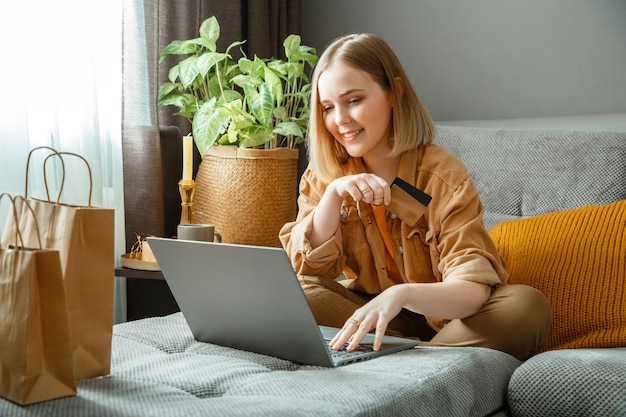 Comprador de mulher jovem e bonita faz compras online usando laptop e cartões de débito de crédito enquanto está sentado no sofá em casa. Comprador de mulher feliz e sorridente adquire bens ou serviços online na sala de estar.