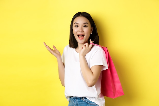Comprador femenino asiático alegre que parece divertido, sosteniendo el bolso de compras y colocándose sobre amarillo.