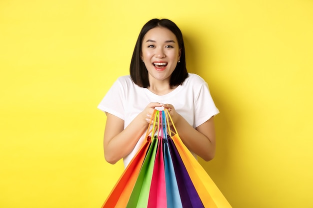 Comprador femenino asiático feliz sonriendo y sosteniendo coloridos bolsos de compras, oponiéndose al amarillo.