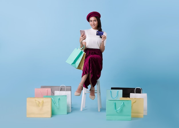 Comprador de hermosa mujer asiática sentado y llevando bolsas de compras con tarjeta de crédito y teléfono móvil en manos sobre fondo azul.