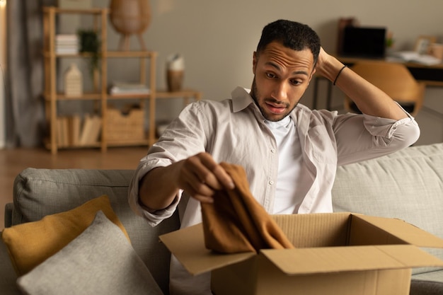 Foto comprador insatisfeito homem negro frustrado desembalando o pacote após a entrega segurando roupas sentadas no sofá em casa