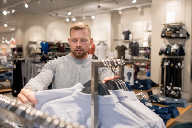 Comprador joven guapo mirando la camisa blanca en el estante mientras elige uno para probarse durante el período de venta en el centro comercial