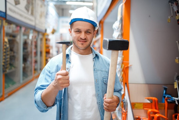 Comprador masculino no capacete branco segurando dois martelos na loja de ferragens.